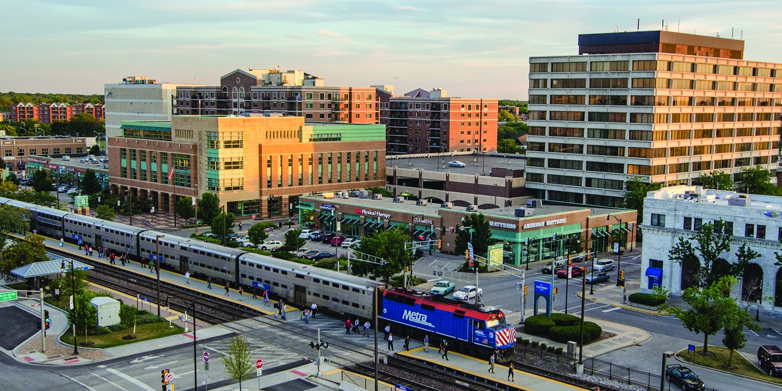 A Metra commuter train stops at the downtown Des Plaines station, surrounded by shops, apartments, and office buildings.