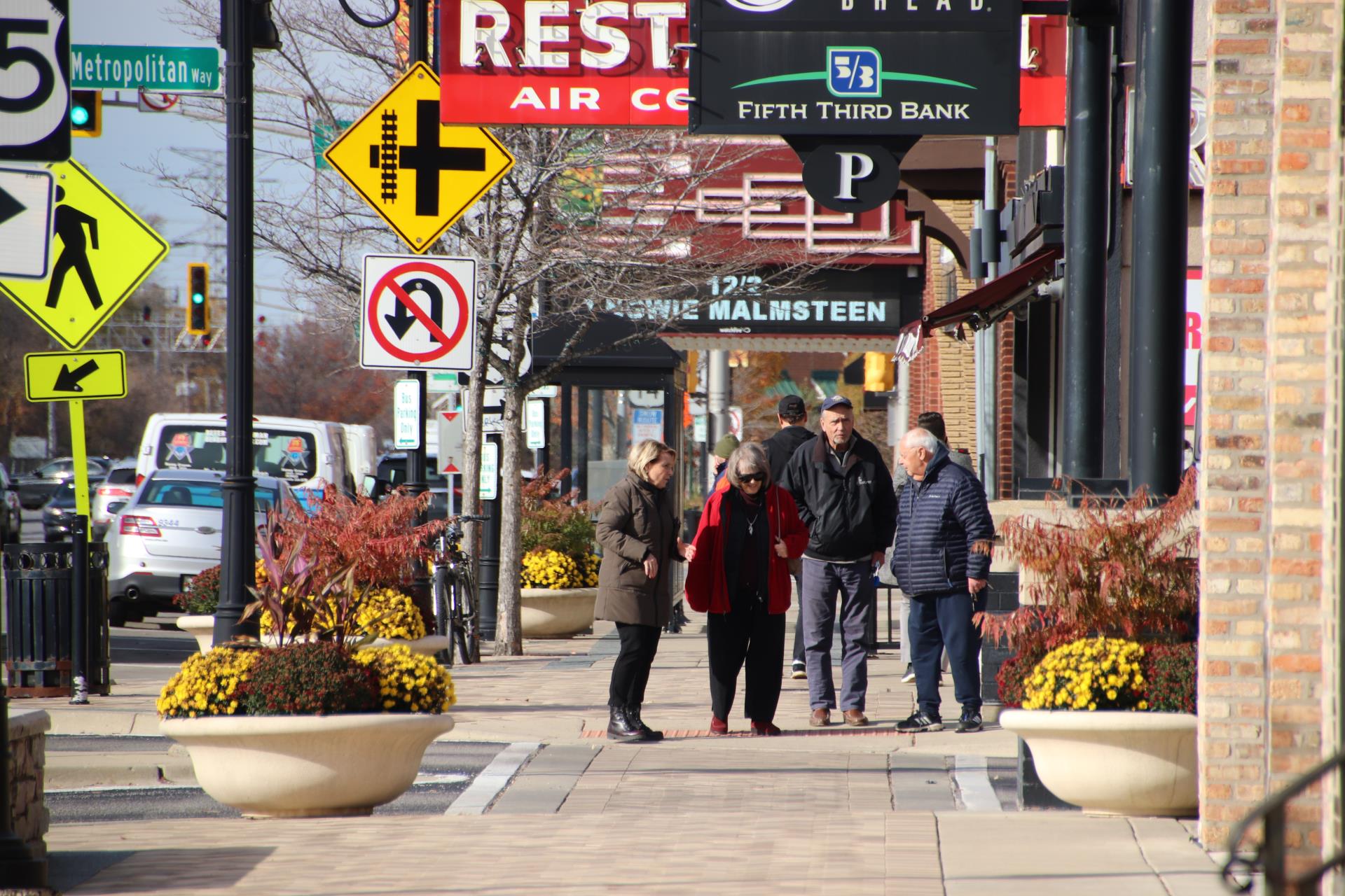 Pedestrians stop to talk on a downtown Des Plaines sidewalk lined with fall planters, storefront signs, and a theater marquee.