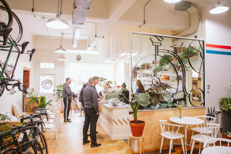 Customers at The Nitro Bar counter inside a bicycle shop, with bicycles, plants, and cafe seating.